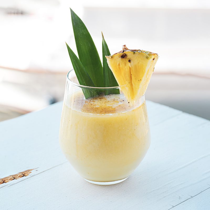 An enticing image of the Pineapple Bliss cocktail, featuring a beautiful yellow drink adorned with pineapple leaves and a slice. Crafted with 1 1/4 oz White rum, 1 1/4 oz Amber rum, and 2 1/2 oz Del Monte Pineapple nectar, this refreshing concoction promises a tropical escape with every sip. The perfect blend of flavors, served over ice, creating a visually appealing and delicious beverage.